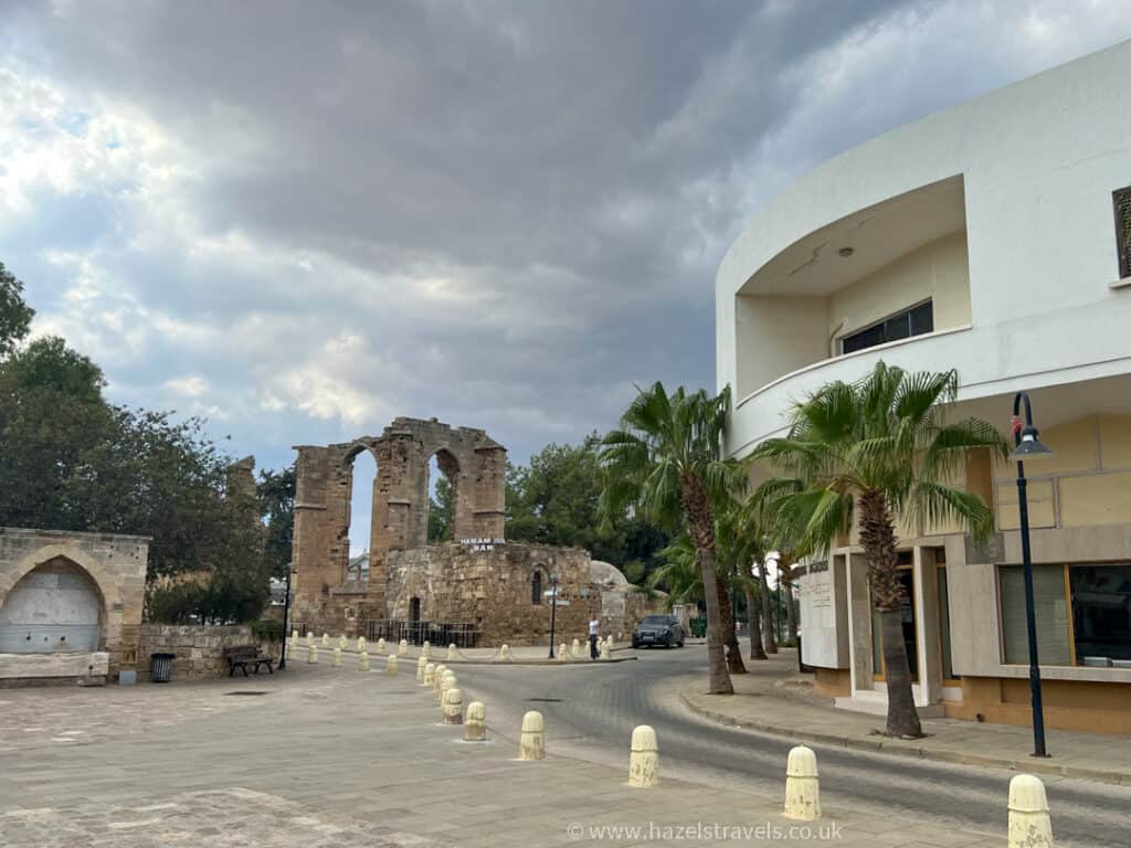 A quiet street curves past ancient stone ruins in Famagusta, with a white modern building, palm trees, and cloudy skies overhead.