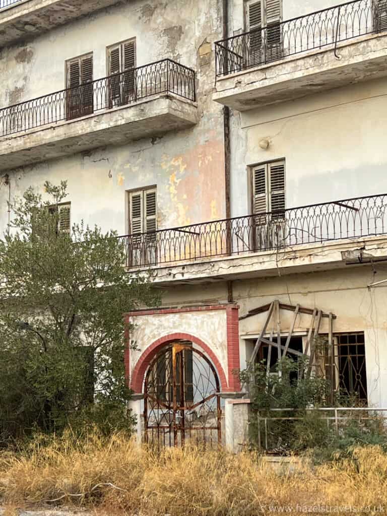 A decaying, abandoned building near the Famagusta border crossing with broken windows, peeling paint, overgrown grass, and rusted balcony railings.