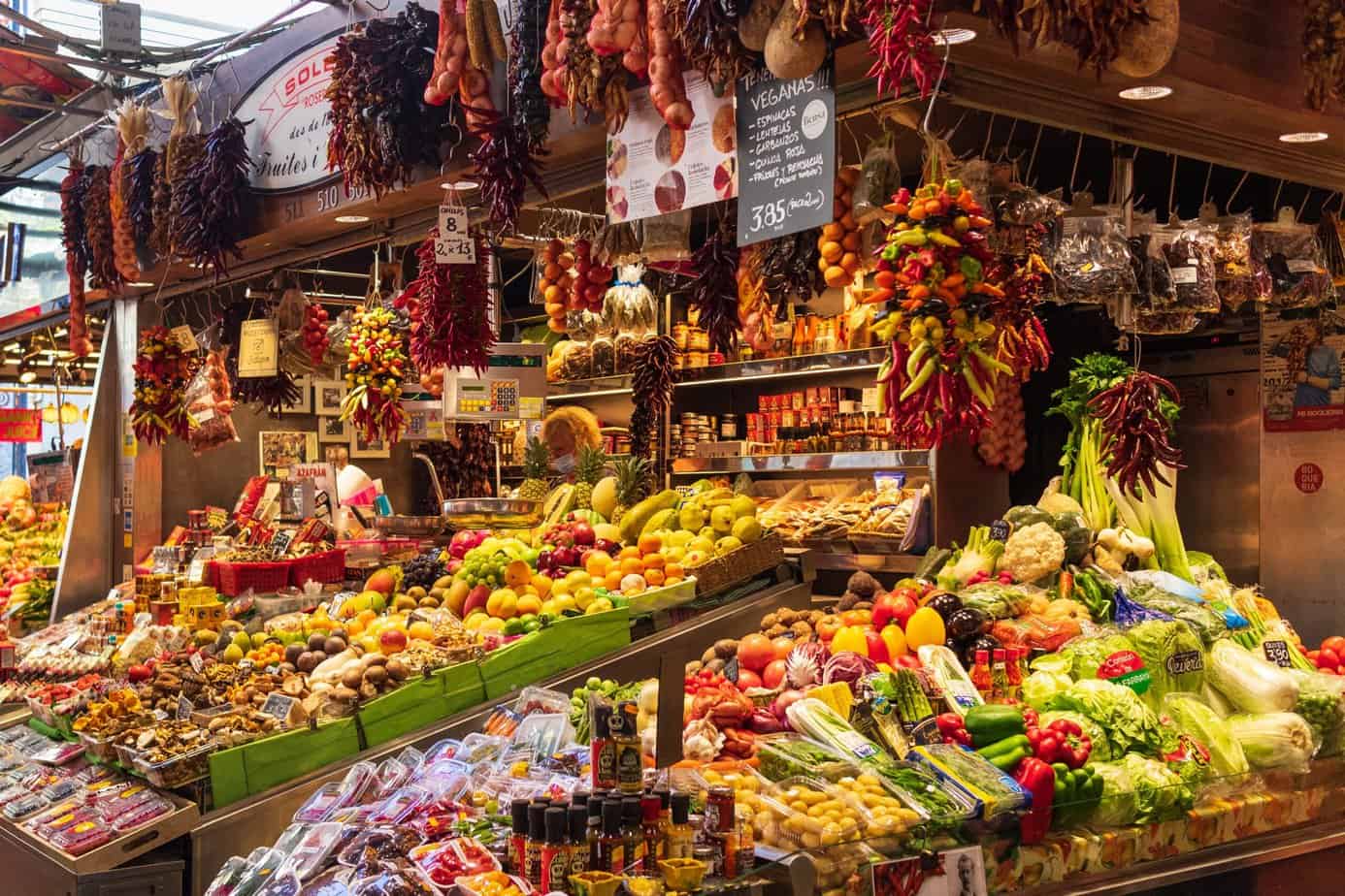 Market stall selling food in Europe, brimming with fresh fruits, vegetables, spices, and cured meats. Bunches of dried peppers and garlic hang above the colourful produce.