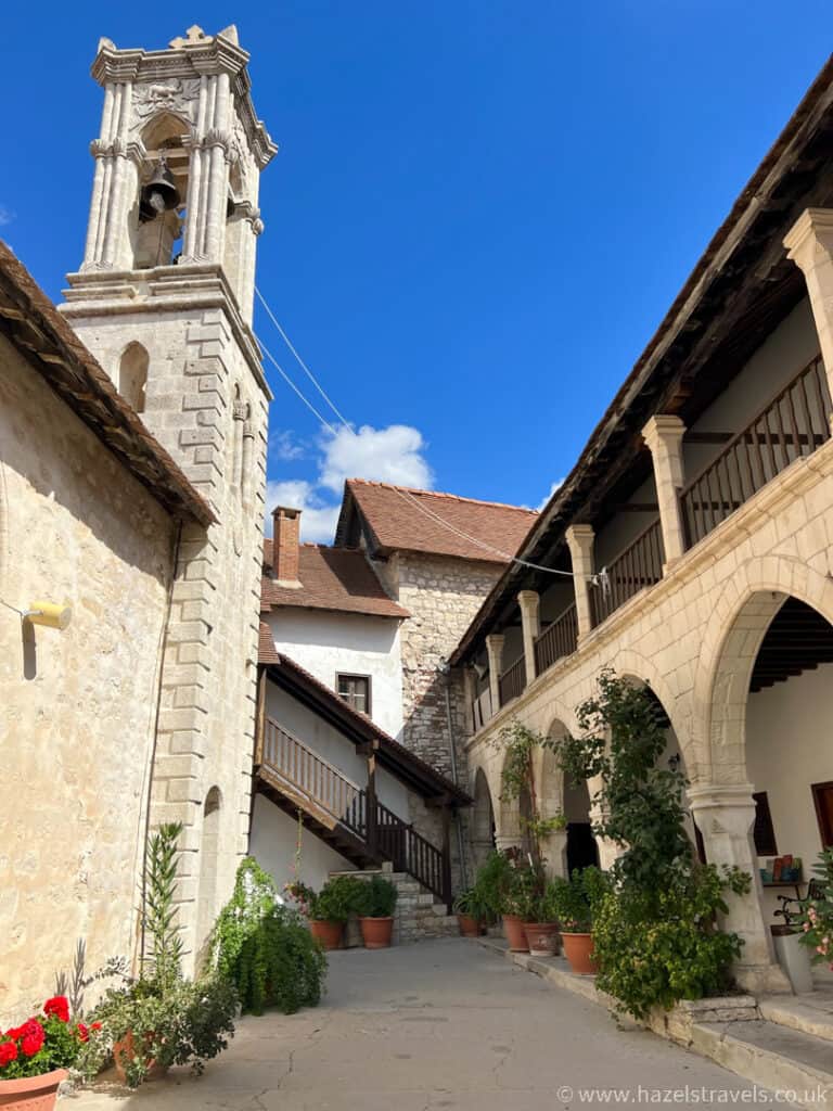 Stone courtyard with potted plants, arched walkway, and a tall bell tower under a clear blue sky.