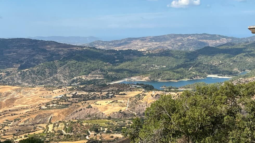 A scenic landscape of the Troodos Mountains in Cyprus featuring rolling hills, patches of green forest, a small lake, and a partly cloudy sky
