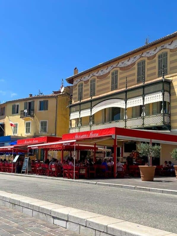 Outdoor café with red awnings and tables in a sunny square in Cassis, France, surrounded by pastel-coloured buildings with traditional shutters.