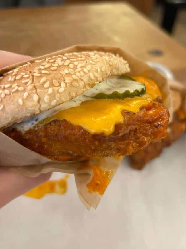 Close-up of a hand holding a fried chicken sandwich with melted cheese, pickles, and a sesame seed bun.