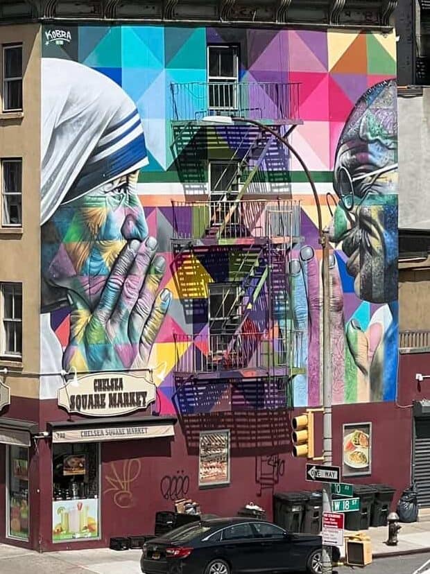 Colorful mural on a building showing two people with hands in prayer, separated by a fire escape. The background is a geometric pattern.