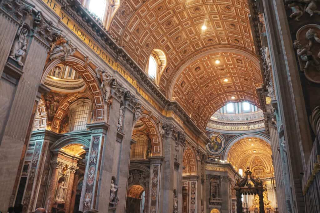 Ornate interior of St Peter’s Basilica in Vatican City, showing grand arches, marble columns, golden ceilings, and detailed sculptures bathed in warm light.