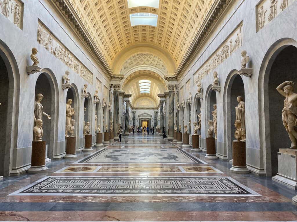 Long marble corridor inside the Vatican Museums, lined with classical sculptures and ornate archways on both sides. The arched ceiling is intricately designed with golden tones, and natural light filters in from skylights, illuminating the polished floors and ancient artwork.