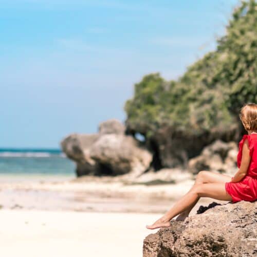 A person in a red outfit sits on a rock at a sandy beach, pondering travel questions and answers while gazing toward the ocean, with trees and rocks in the background.