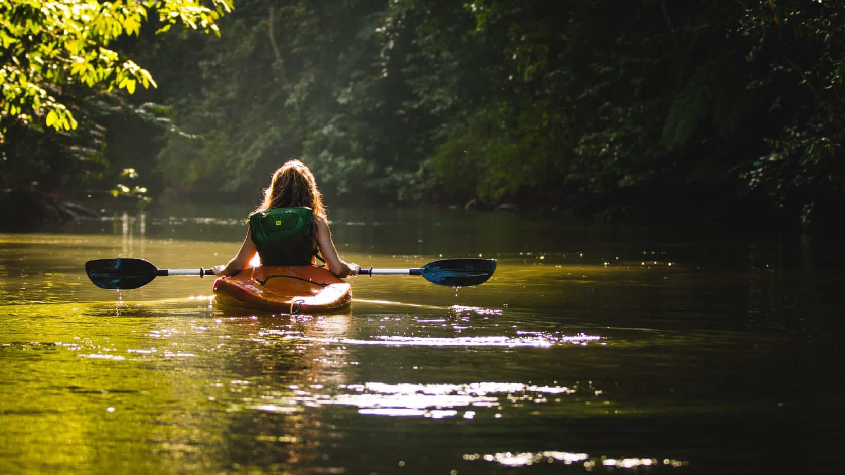 A person with long hair kayaks alone on a calm river, discovering the benefits of travelling alone. They glide through the serenity, surrounded by dense green foliage under dappled sunlight.