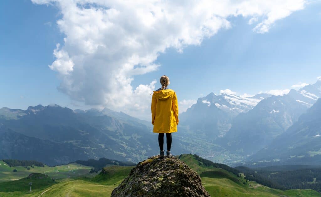 A solo female traveller in a yellow coat stands on top of a rock and looks out over mountain peaks and clouds