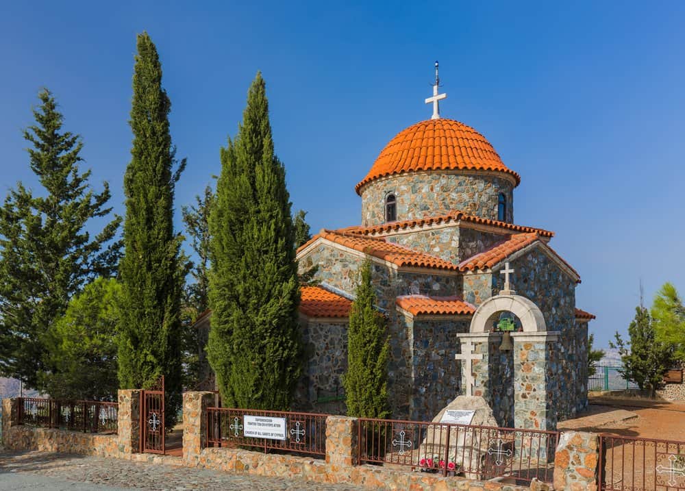 Stone church with a red-tiled dome and cross, surrounded by tall cypress trees and a metal fence, set against a clear blue sky