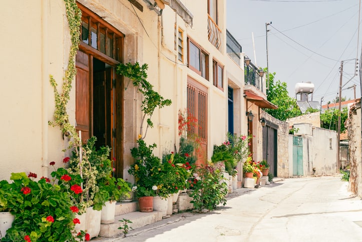 A narrow sunlit street in Cyprus lined with houses featuring wooden doors and windows, with potted plants and flowers arranged along the walls