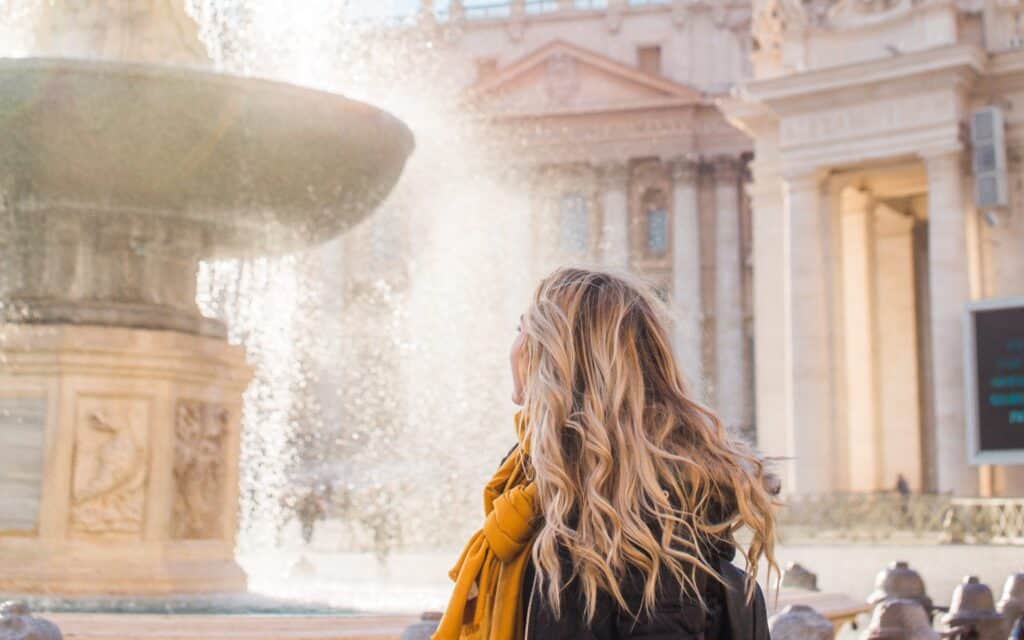A female traveller with long blonde hair and a yellow scarf faces a large, ornate fountain in a sunlit, classical architectural setting.