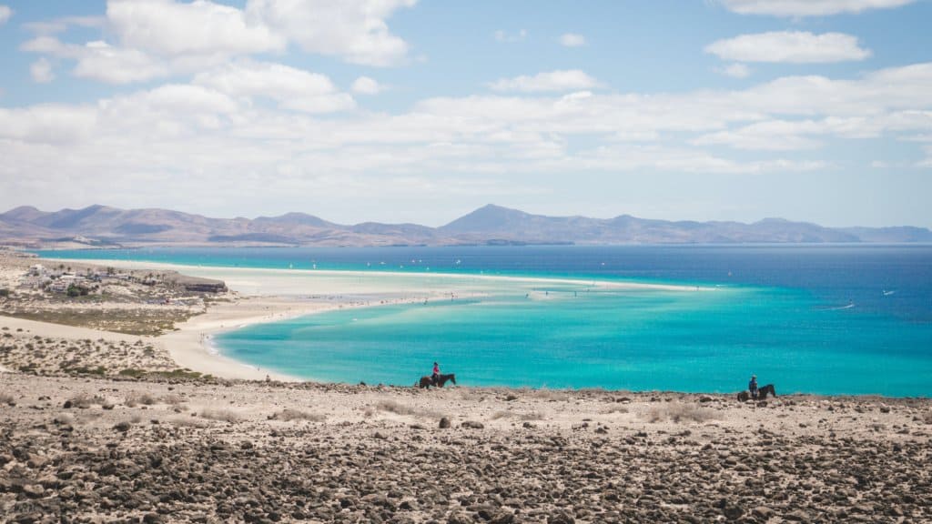A wide sandy beach curves along turquoise water under a partly cloudy sky, with two people riding horses on a rocky foreground. Hills and mountains are visible in the background.