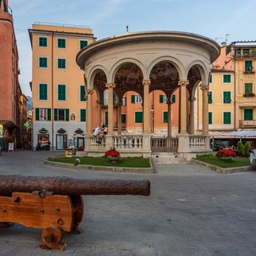 Historic square with a rusted cannon on a wooden platform, in front of a circular pavilion with arches. Surrounding are colorful buildings with green shutters and outdoor seating.