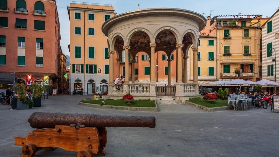 Historic square with a rusted cannon on a wooden platform, in front of a circular pavilion with arches. Surrounding are colorful buildings with green shutters and outdoor seating.