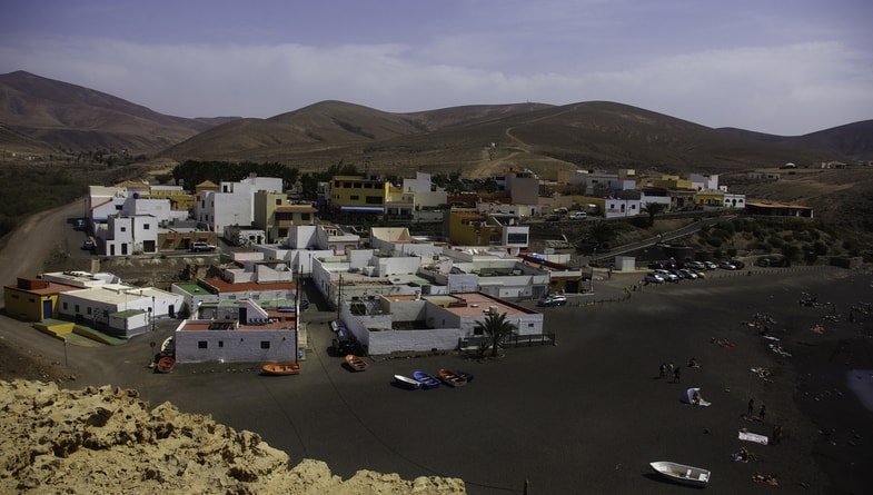 Small coastal village with flat-roofed buildings, surrounded by brown hills, cars parked along the road, and people on a dark sandy beach with small boats.