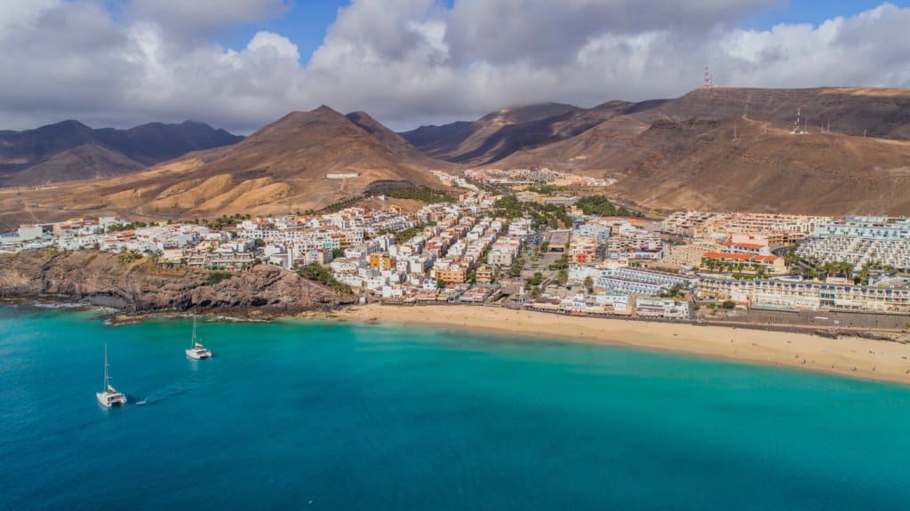 Aerial view of a coastal town with white buildings, sandy beach, turquoise water, anchored boats, and mountains in the background under cloudy skies.