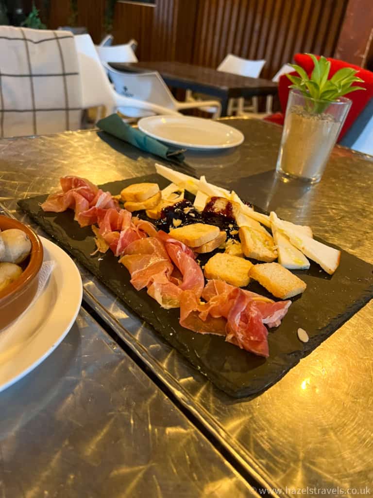 A slate platter with sliced cured meat, cheese, crostini, and jam is served on a metal table in a casual restaurant setting.