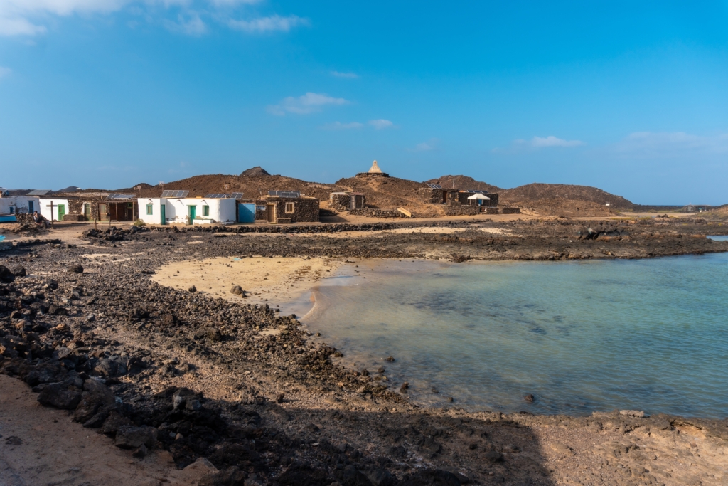 Small coastal village with simple buildings near a rocky shoreline and clear blue water, set against arid hills under a bright sky, reminiscent of Lobos Island Fuerteventura.