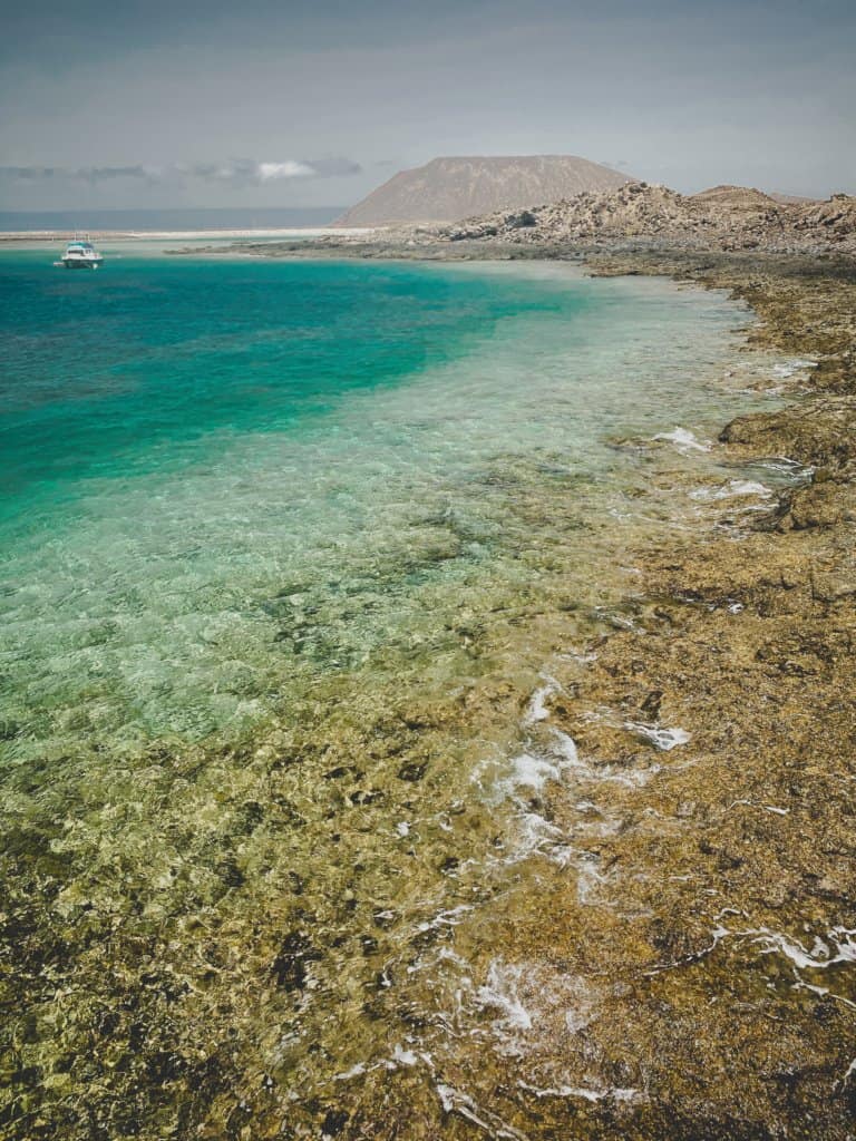 Coastal view of crystal clear waters flowing over brown rocks, volcano in the distance. Green water and one white boat.