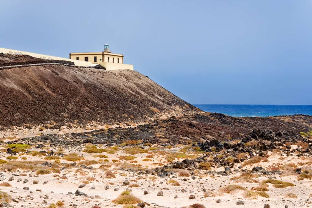 Brown Sandy beach with yellow sand and brown rocks, with a square cream coloured lighthouse on top of a hill and blue sea.