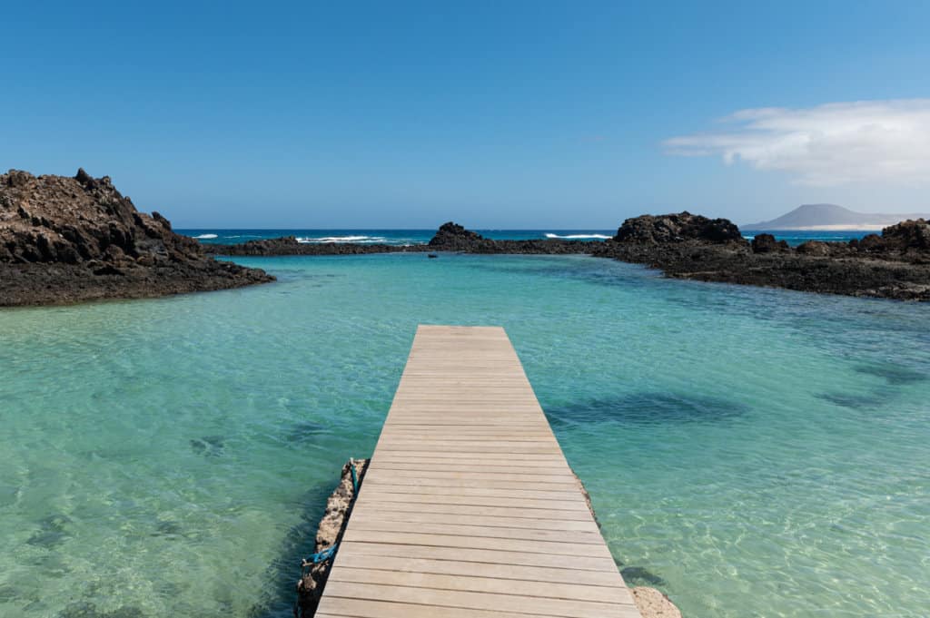 Wooden Pier stretches out into crystal clear blue and green waters, with black volcanic rocks around the edges and a blue sky