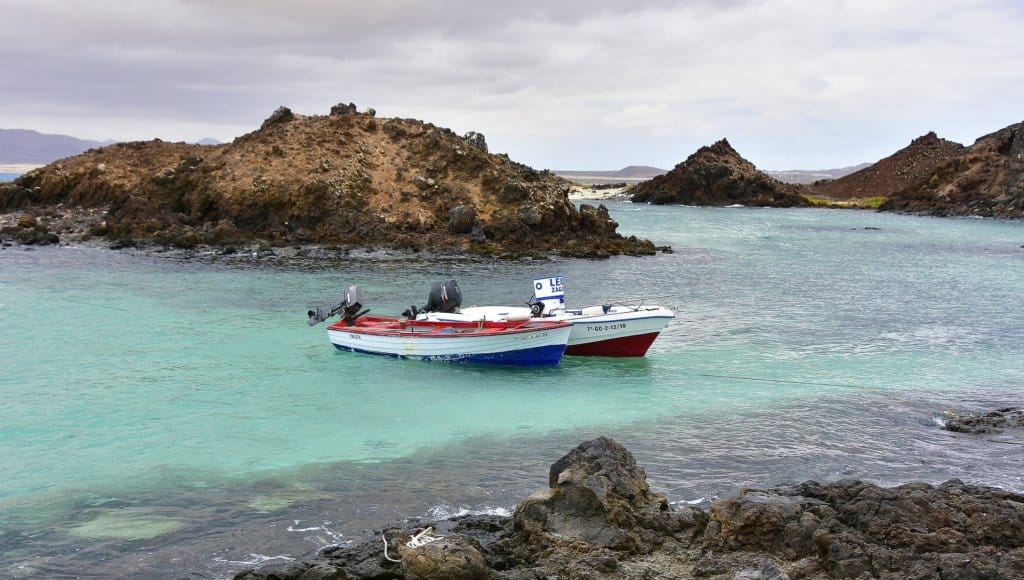2 small boats in the shallow green waters of Lobos Island, with volcanic cones and mounds protruding from the water. Cloudy skies.