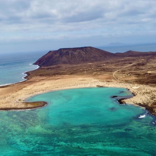 Aerial view of Lobos Island with blue/green water, crescent shaped yellow sand beach and large brown volcano caldera in the distance.