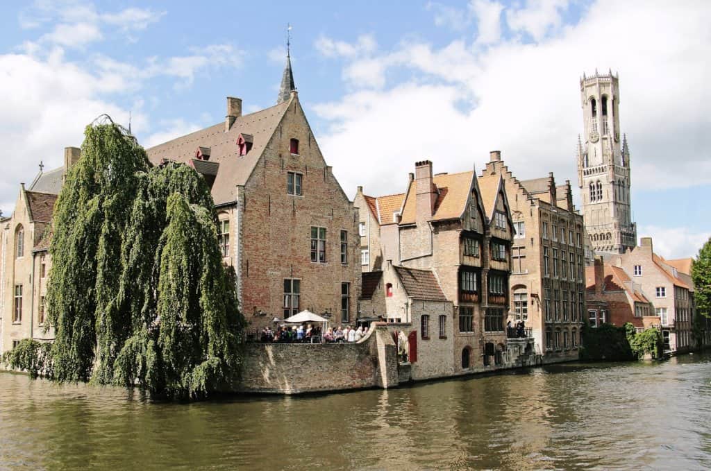 Brown river view with brown brick four storey buildings and a tall circular tower. Green tree can be seen and people sitting at tables and chairs outside the largest building.
