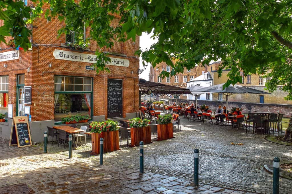 Belgian red-bricked brasserie with cobblestone outdoor area and red tables and chairs with people sitting for lunch. Green canal can be seen in the distance. Potted planters with red flower. Chalkboard on the wall. Green leaves from a tree in the foreground.