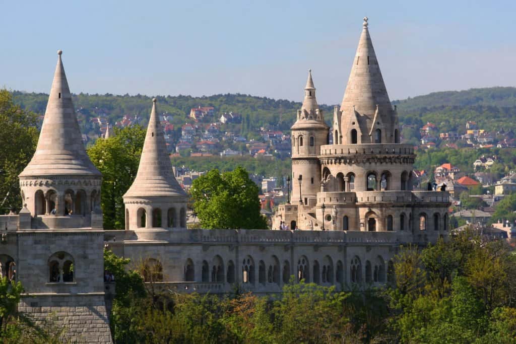 Romantic style castle with pointed spires in a cream coloured stone. Green hilltops with orange rooftops in the distance.