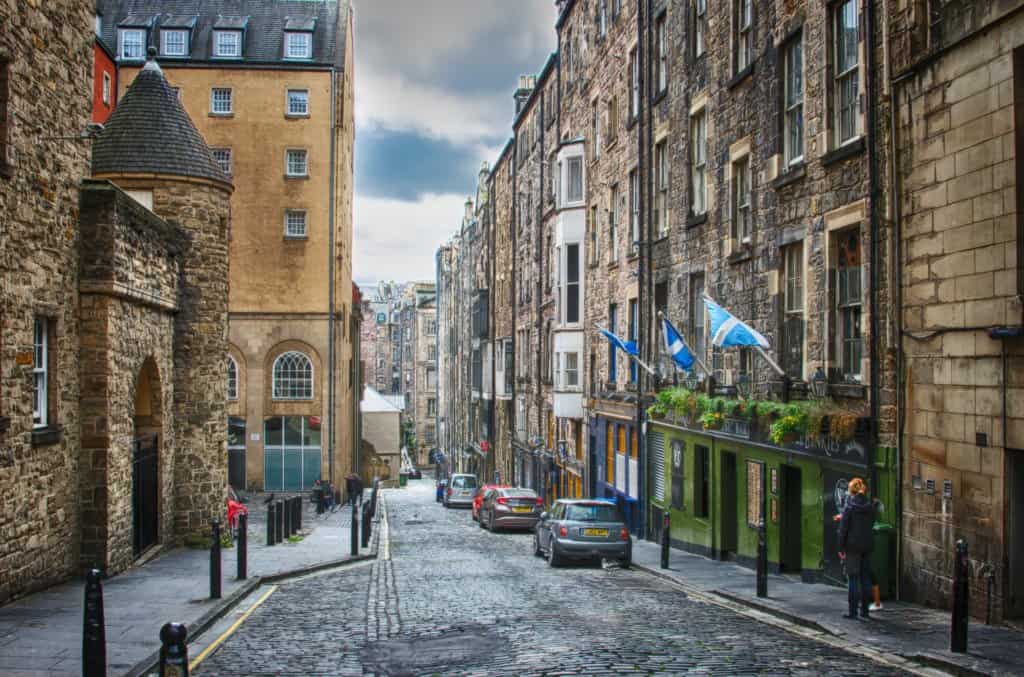 Narrow cobbled street with tall grey buildings on both sides. Cobbled road down the centre with parked cars. Green shop front on the right with three blue and white Scotland flags.