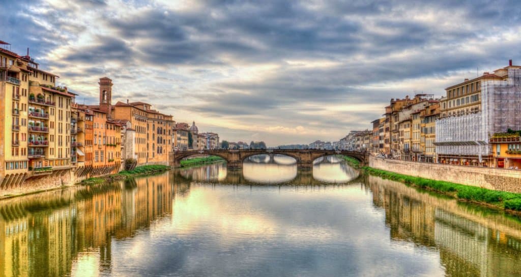 Landscape view of a calm river at golden hour. Orange and yellow buildings line the banks and reflect in the calm water. A brown bridge across the river is the main focal point. Blue and grey clouds in the sky with accents of yellow and pink.