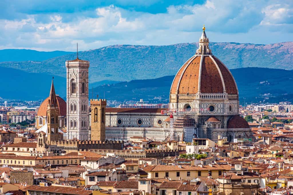 Beige and brown coloured city view from above, terracotta rooftops in the foreground with a huge terracotta dome in the centre on a large renaissace style building or castle. Green mountains in the background, blue sky.