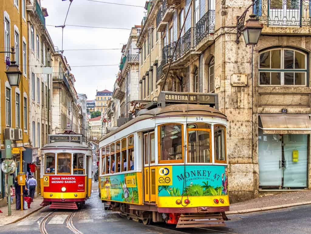City street view with 2 trams central to the photo with tall beige coloured buildings either side. Yellow tram and red tram, Portuguese writing, grey cloudy sky.