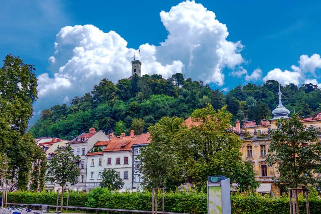 Landscape view of a town in Europe during the day. Bright blue sky, white clouds, coloured buildings in orange and blue in a terraced row, green hedge and trees in the foreground.
