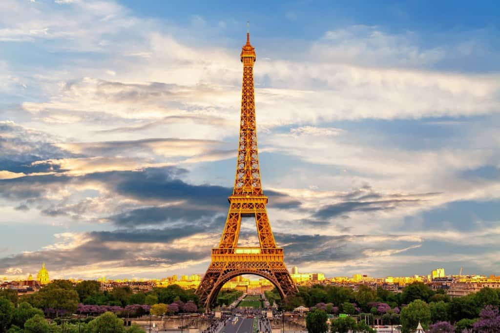 Landscape view of the Eiffel tower during golden hour. Blue sky with grey clouds. Lit up orange buildings on the horizon. Green trees and foliage in the foreground. Eiffel tower itself an orange hue.