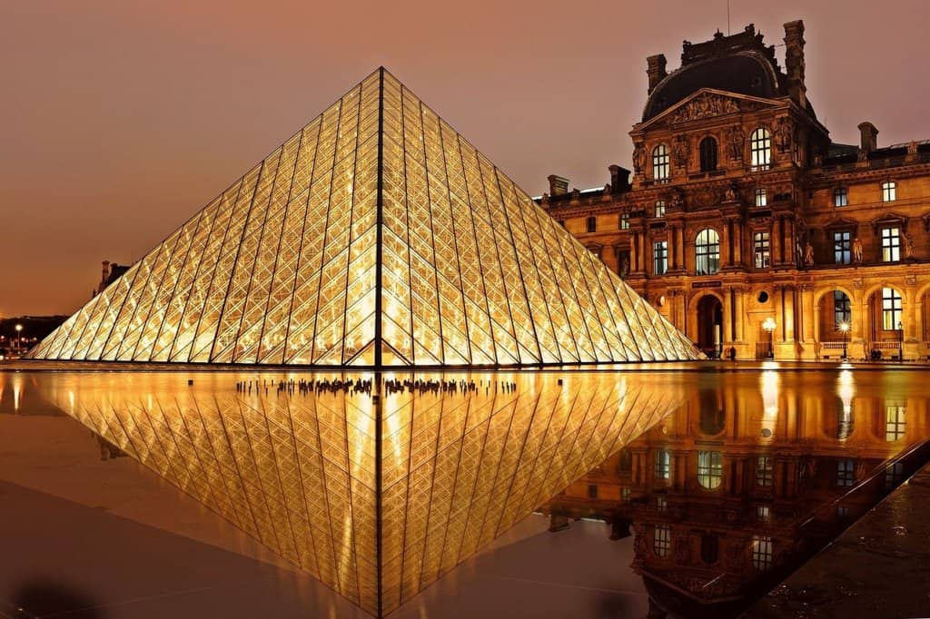 Sunset view of the Louvre in Paris. Triangular glass sculpture lit up in yellow and reflected in water on the ground. Orange buildiing in the background, pink sky, noo people.