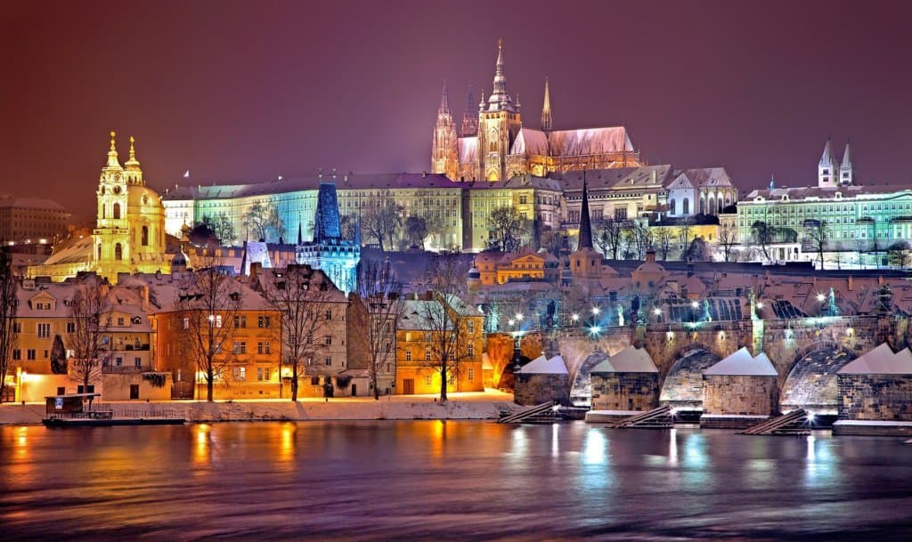 Pretty picture postcard scene depicting a European city (Prague) lit up in various pastel colours. View from over a river, with a castle with spires on top of a hill in the distance.