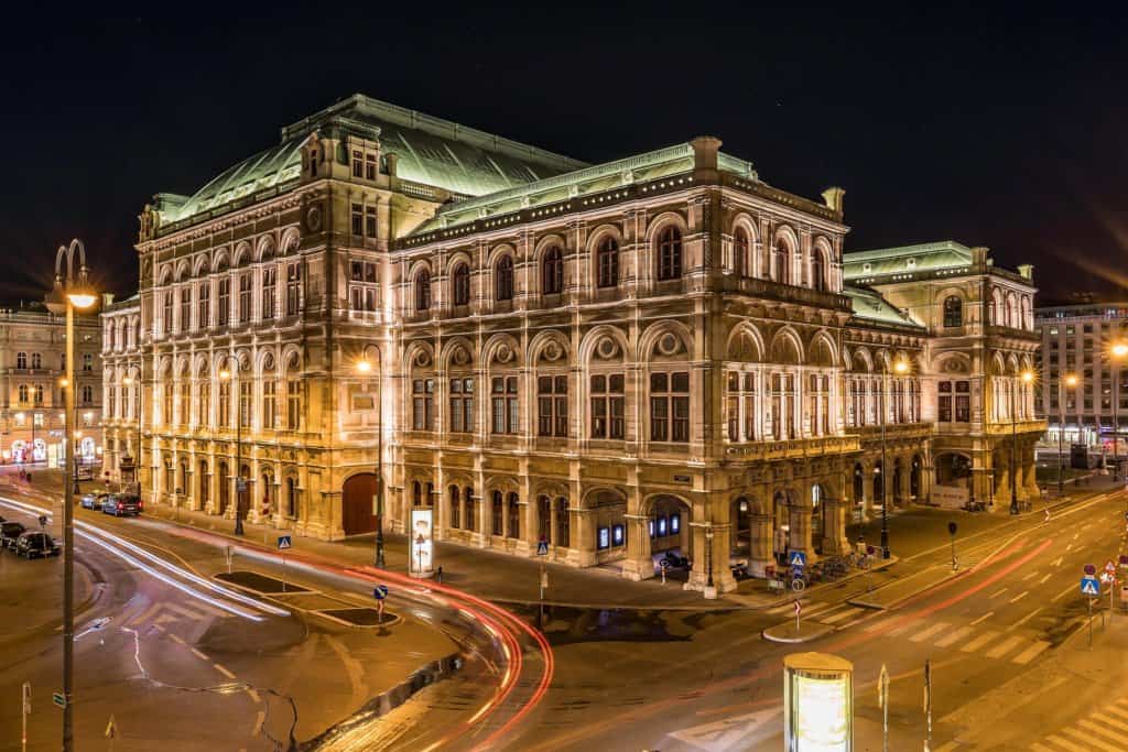 The oranate renaissance building of Vienna State opera at night, with empty roads and streets with street lights and some red traffic trails.