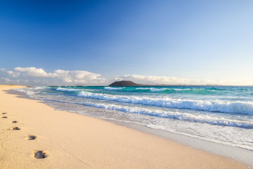 A beach scene, yellow sand with footprints, blue sea with waves and blue sky with white clouds. Small volcanic island (Lobos Island) on the horizon.