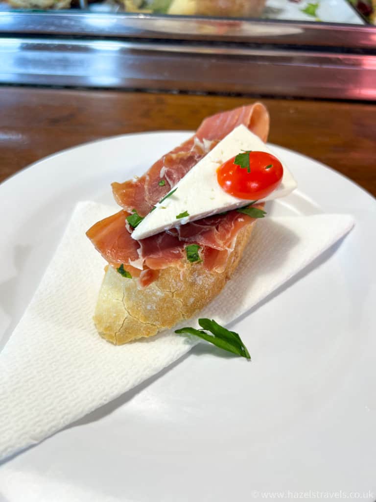 A Spanish pinxto - slice of bread with parma ham, white cheese and slice of tomato - on a white napkin on a white plate.