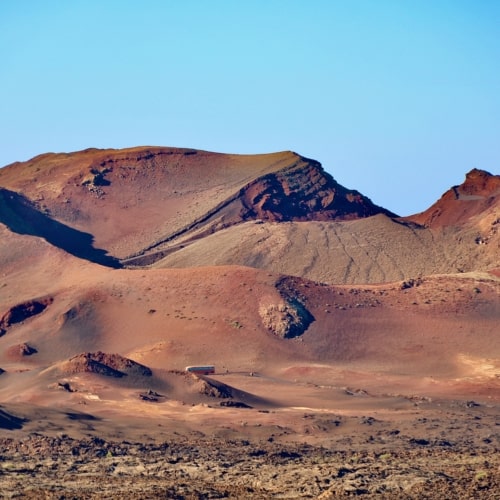 Huge red and brown volcanic mountains rising up into a bright blue sky. Two tiny buses can be seen in the foreground driving along the road to the crater of Mount Timanfaya in Lanzarote.