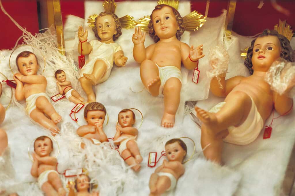 Assortment of baby Jesus figurines with labels, displayed on white fabric.