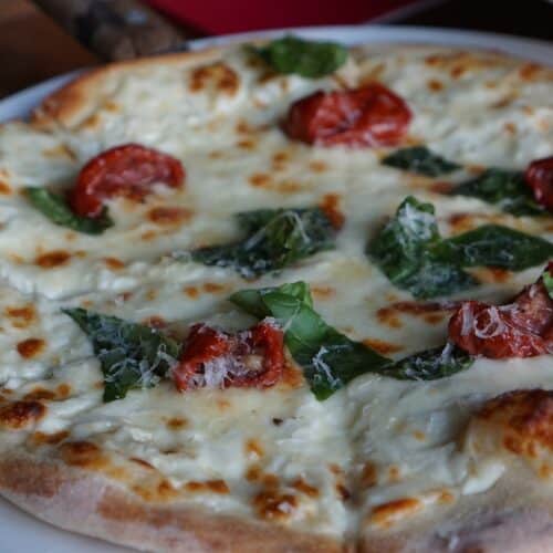 A freshly baked pizza topped with melted cheese, spinach leaves, and roasted tomatoes, served on a white plate.