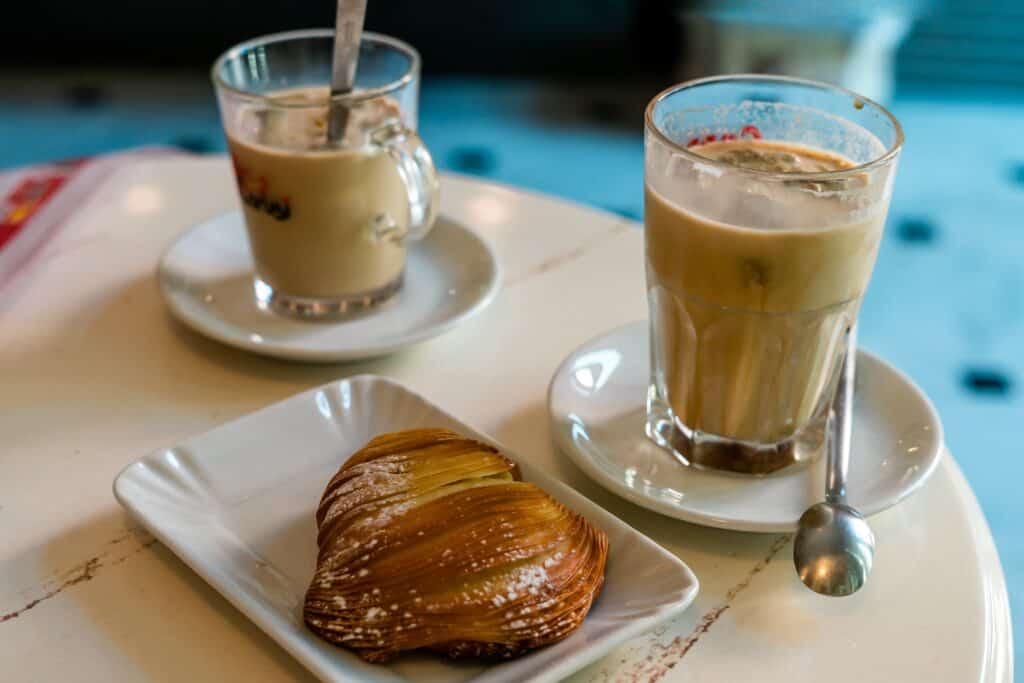 Two glasses of coffee on saucers, one with a spoon, and a pastry on a rectangular plate on a white table.