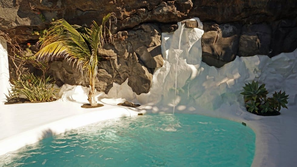 A small green pool surrounded by white painted rocks and brown volcanic rock, green foliage and palms, small white fountain pouring into the pool.
