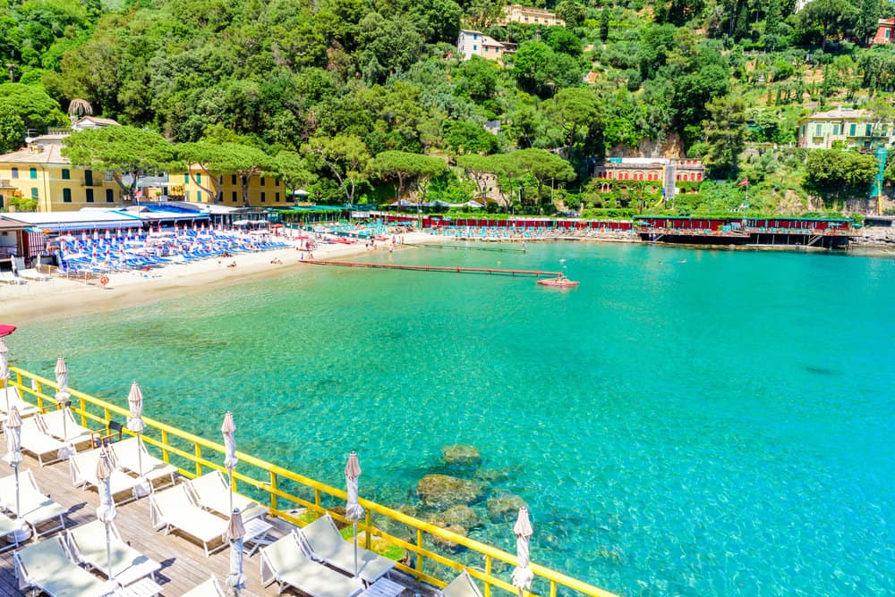 Paraggi Bay Beach near Portofino, Italy — a picturesque cove with crystal-clear turquoise water, golden sand, sun loungers, and colourful beach clubs backed by lush green hills.