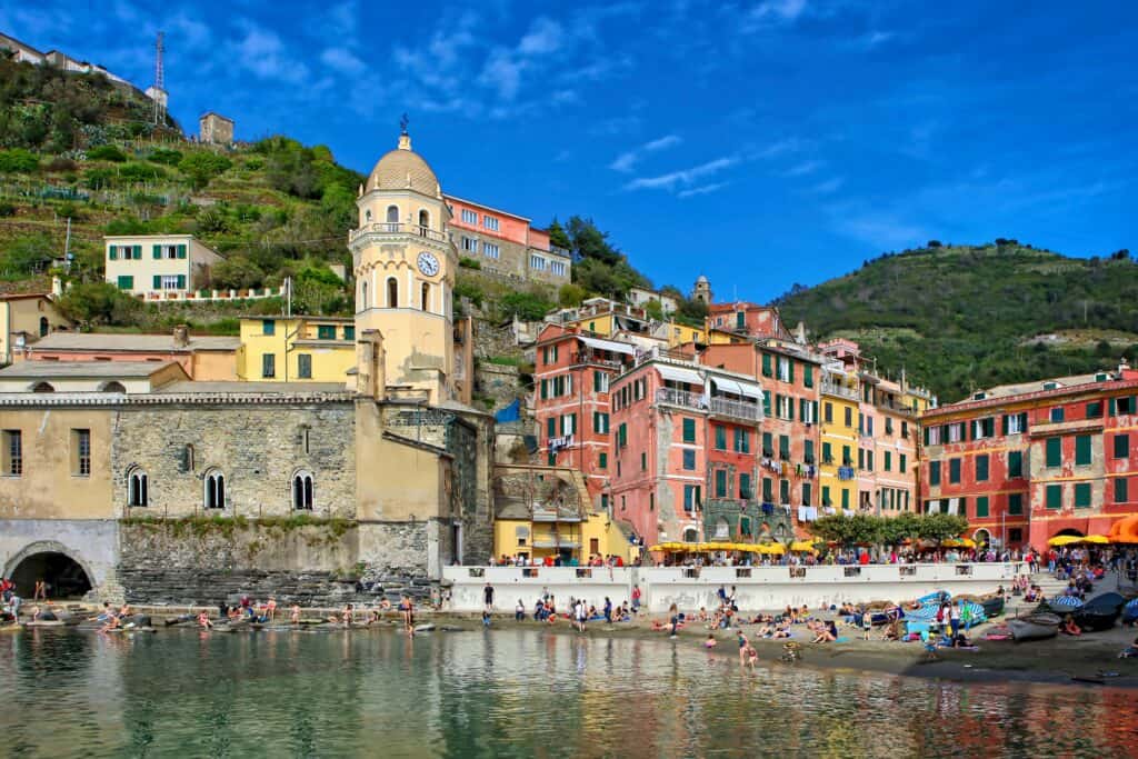 Colorful hillside buildings and a church with a clock tower overlook a busy waterfront with people relaxing by the water.