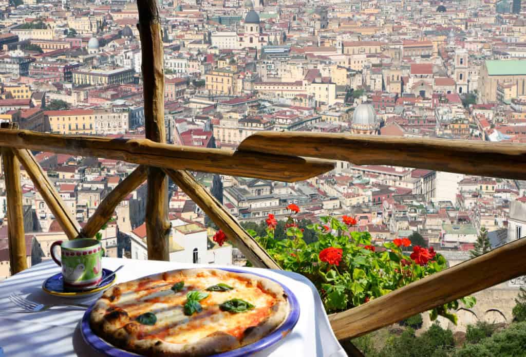 An aerial view of a European city with orange and brown rooftops and decorative domes. Wooden barriers at the top of a balcony with a plate of pizza and a cup of coffee in the foreground on a table with a white tablecloth. Red flowers can be seen jut below the balcony.